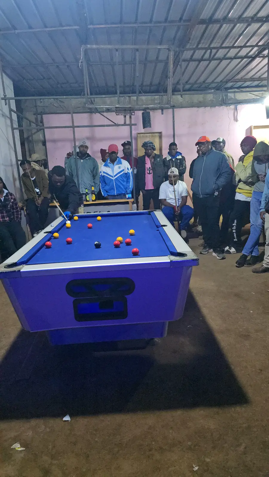 A village pool joint at night: iron-sheet roof, dirt floor, crowd of young men